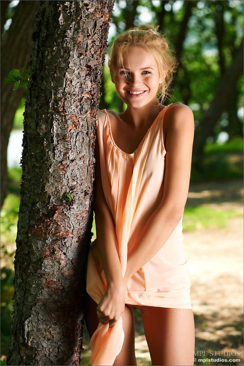 Smiley cute teen in short dress disrobes to pose nude under a tree page 1