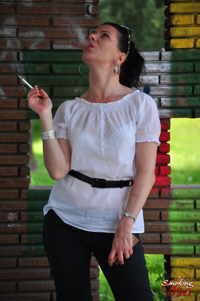 Fully clothed brunette Mina smokes a cigarette in front of a brick wall page 1